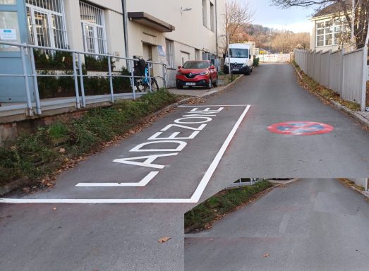 Weiß markierte Ladezone mit Schriftzug „LADEZONE“ und Halteverbotssymbol auf Asphaltstraße neben Gewerbegebäude..