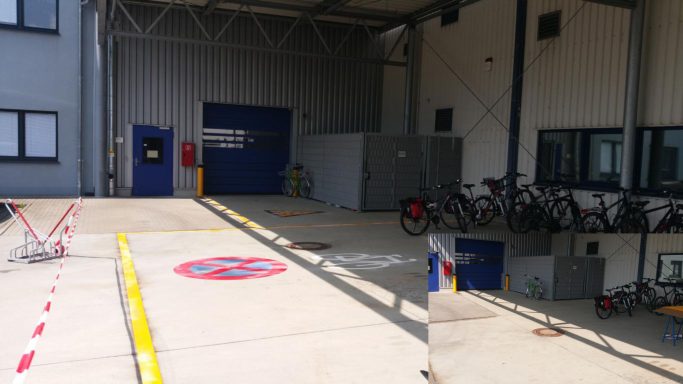 Außenliegender Verkehrsbereich mit gelben Bodenstreifen, Halteverbot-Markierung und Fahrradsymbol vor Industriegebäude